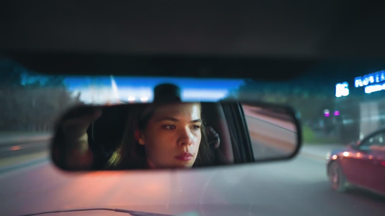 여성은 차를 운전하는 동안 뒷모습 거울에 반사되며, 도로와 그녀의 앞에 있는 빨간 차는 배경에 보이고, 불빛이 켜져 있으며, 희미하게 밝은 저녁을 통해 이동합니다.