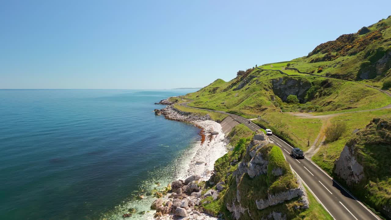 High aerial video of the Causeway Coastal Route in County Antrim, Northern Ireland, UK, on a bright and sunny day. Cars pass along the cliff-side road. Filmed in 4K, 60FPS and with Rec709 color.