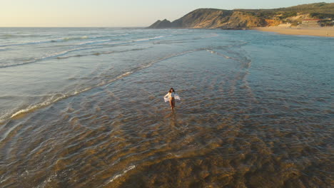 Vista-Aérea-De-Una-Mujer-En-Bikini-Y-Vestido-Blanco-Caminando-Por-La-Playa-Durante-La-Marea-Baja-Durante-La-Puesta-De-Sol.