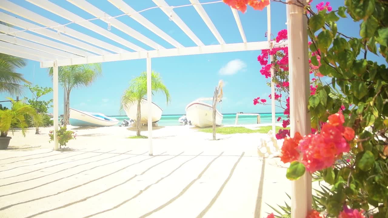wide shot serenity of high-end Caribbean resort beach, framed by a white slatted pergola and bright tropical flora