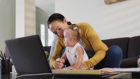 madre caucásica sosteniendo a su bebé tomando notas mientras trabaja desde casa