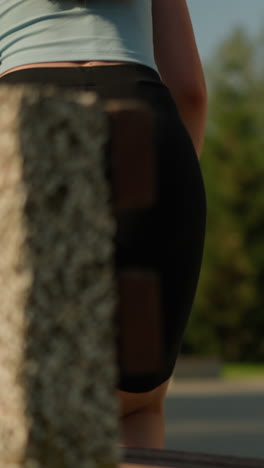 young lady holding cyan roller skates as she drops them on wooden park bench, she wears black tights and light blue top with a blurred background featuring tall trees, greenery, and lamp post