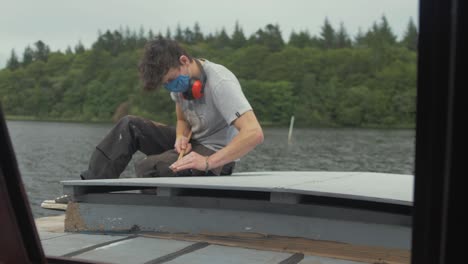 young caucasian man sanding wooden teak trim with sandpaper on boat by river