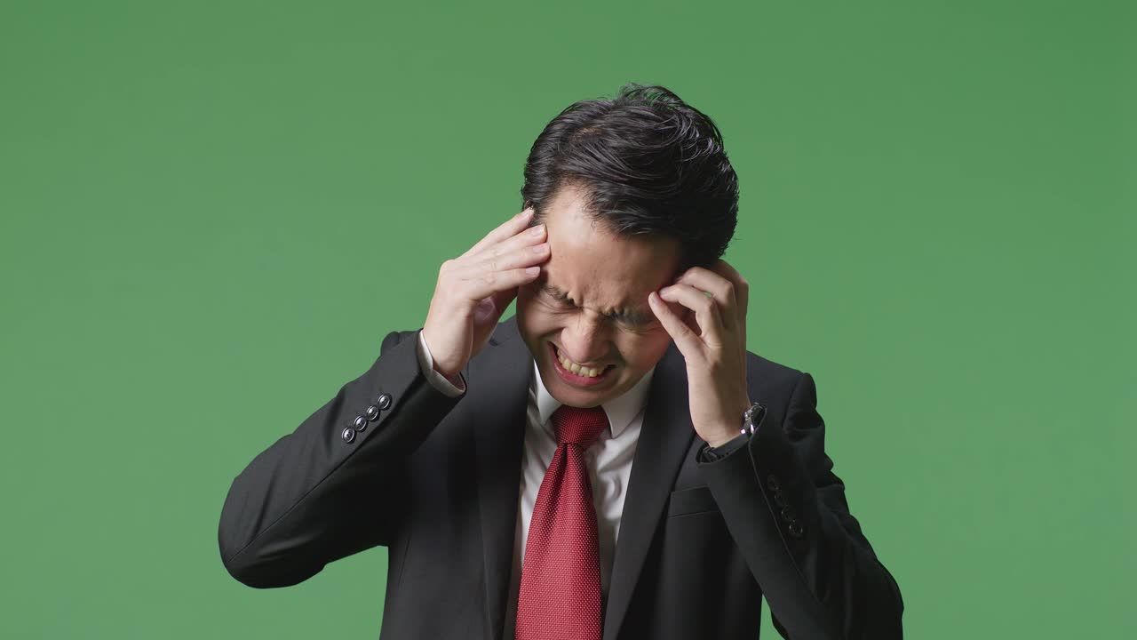 녹색 스크린 앞 에 서 있는 아시아인 사업가 가 두통 을 고 있다