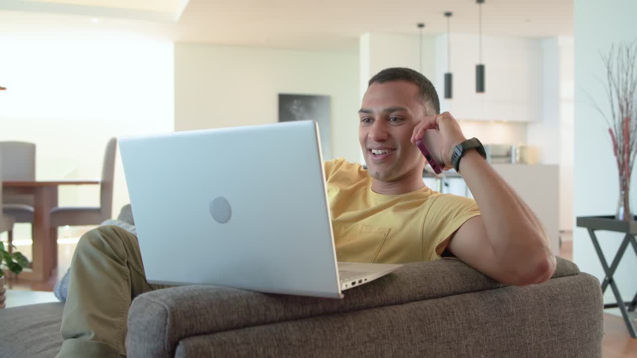Receiving smartphone call, man sitting on gray sofa typing on silver laptop, smiling in a luminous l