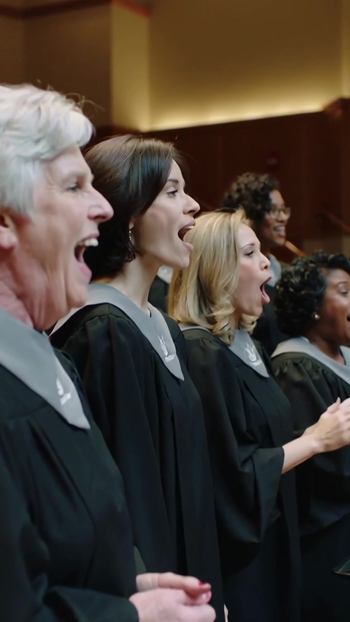 Video verticale: Coro che canta in tuniche nere e stole grigie dopo la nota di apertura su una piattaforma, applaudendo.
