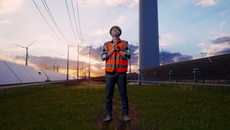 full body of asian male engineer with safety helmet pray for something while standing with solar panel and wind turbines