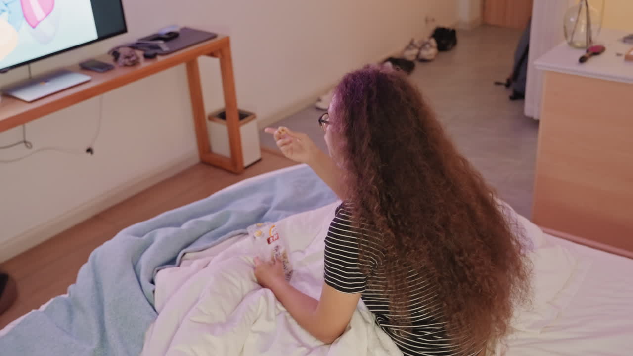 Jonge vrouw op hotelbed kijkt tv, pakt de afstandsbediening en past de zender aan. Lang krullend haar valt over het dekbed, zachte deken en kussen, minimalistische houten console met gloeiend scherm, pantoffels