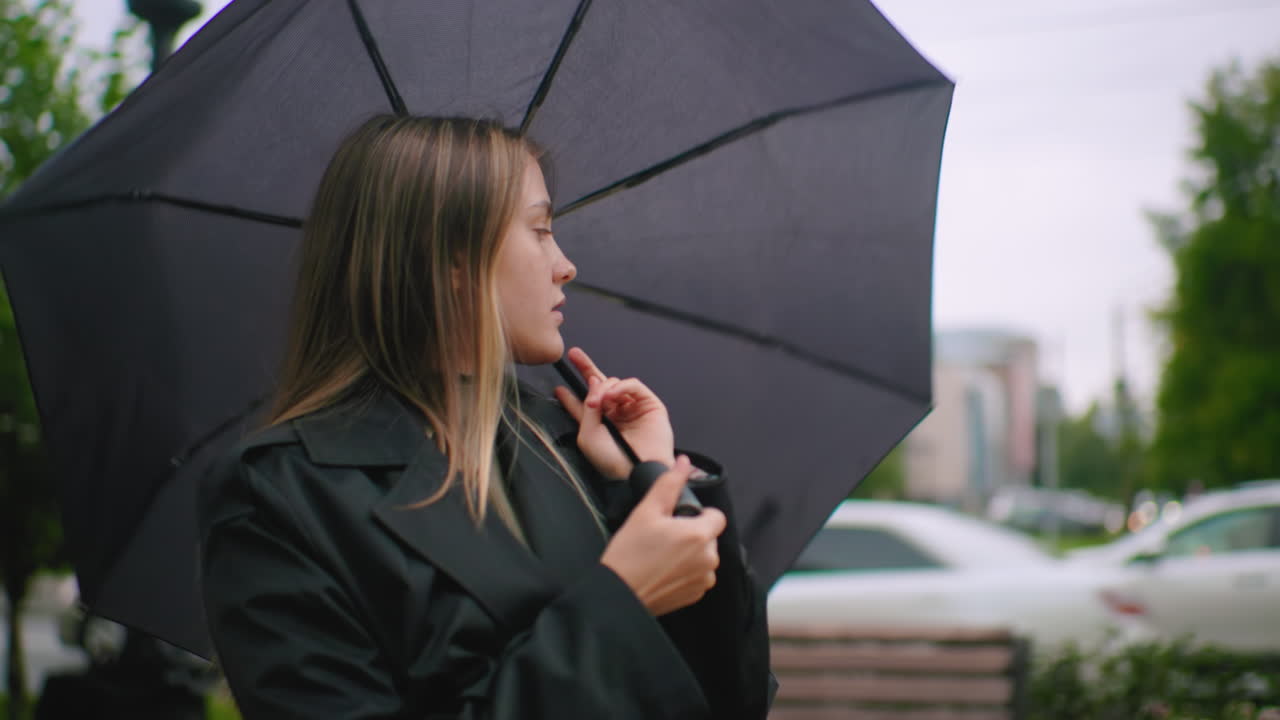 Gentlewoman in black coat spinning umbrella looks into distance on rainy windy city street with blurred cars and greenery in background, appearing thoughtful and reflective in urban outdoor environment