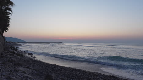 Ein-Zeitraffervideo-Fängt-Den-Atemberaubenden-Sonnenaufgang-über-Dem-Meereshorizont-Ein,-Wie-Er-Von-Der-Steinigen-Küste-Aus-Gesehen-Wird