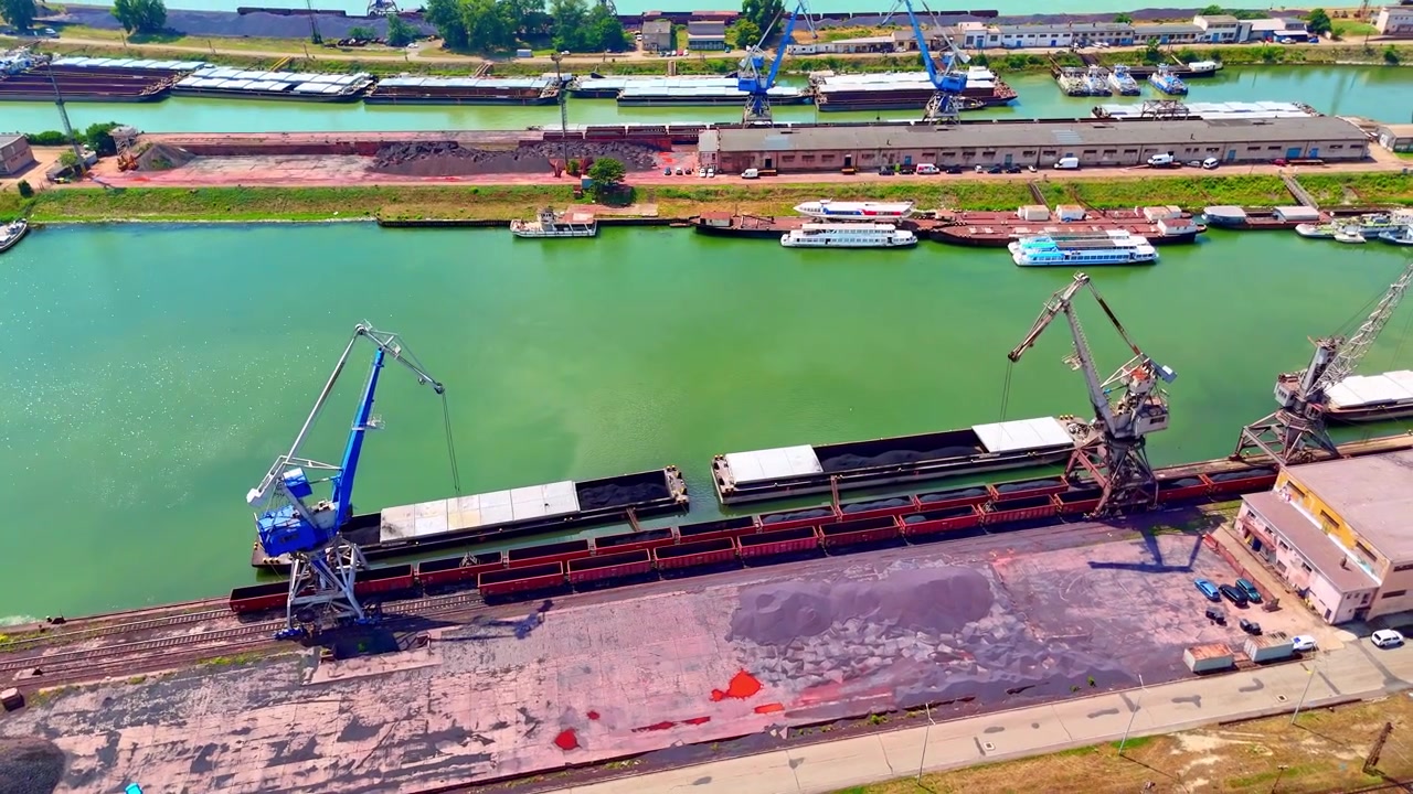 Chiatte da carico nel porto fluviale industriale, Bratislava, Slovacchia. Prospettiva drone di gru di carico blu e lunghe imbarcazioni sull'acqua verde in pieno giorno.