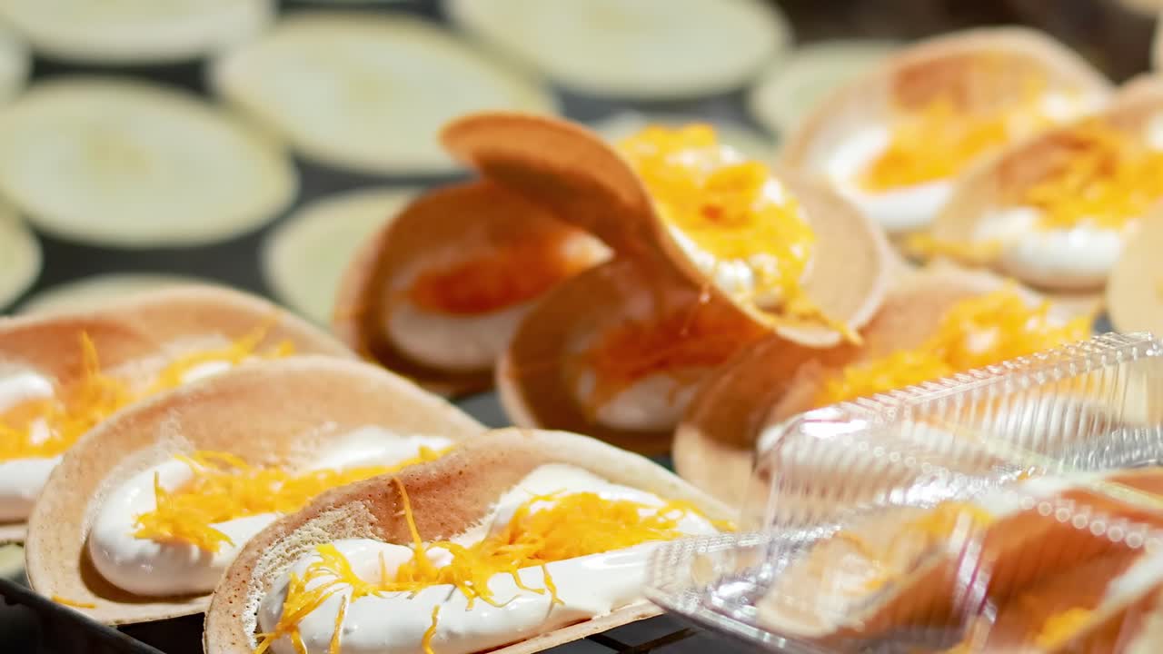 Detailed view of Thai pancakes topped with cream and golden threads in a plastic container.