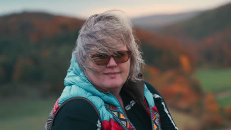 Primer-Plano-De-Una-Mujer-Gorda-Sonriente-Con-Pelo-Gris,-Gafas-Y-Chaleco-Cálido-Y-Colorido,-Rodeada-De-Una-Hermosa-Naturaleza-Otoñal-Con-Naranjos-Al-Fondo-Durante-Una-Puesta-De-Sol-Fría-Y-Ventosa