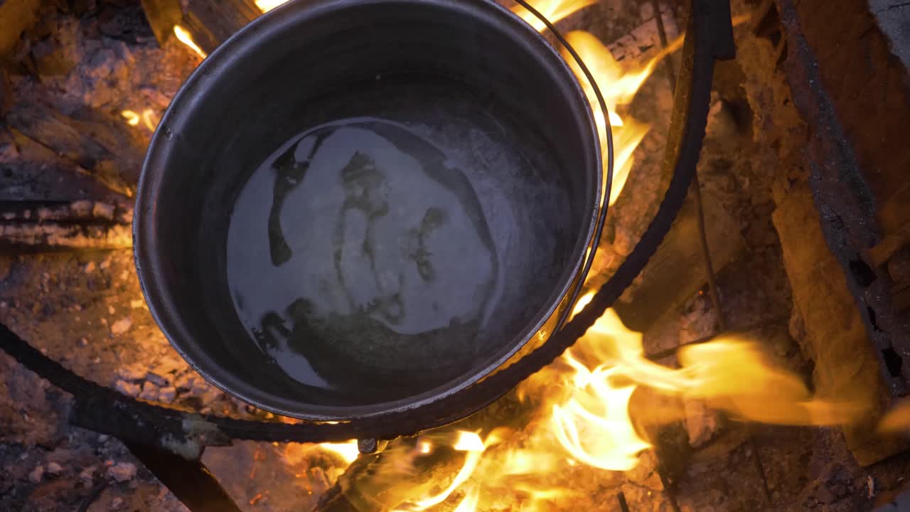 Black cauldron burning on the fire in nature near a cottage. The cauldron has oil that sizzles slightly on the fire. The fire is very intense and bright red