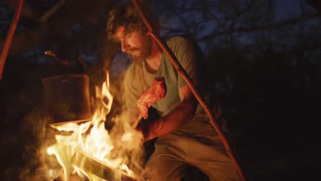 bearded caucasian male survivalist in wilderness cooking meat on stick and heating pot in campfire