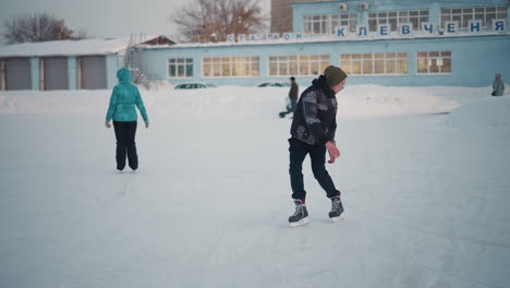 skillful boy skating with ease on snowy ice rink surrounded by other skaters and onlookers near urban buildings under pale winter sky capturing smooth motion and seasonal leisure atmosphere