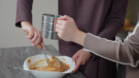 partial view of girl helping her aunt in kitchen, holding metal sifter while aunt stirs batter in bow, they work together in bright, modern kitchen