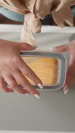 young housewife shakes tall plastic container looking at movement of spaghetti. woman closes package with lid stocking raw food in kitchen upper view