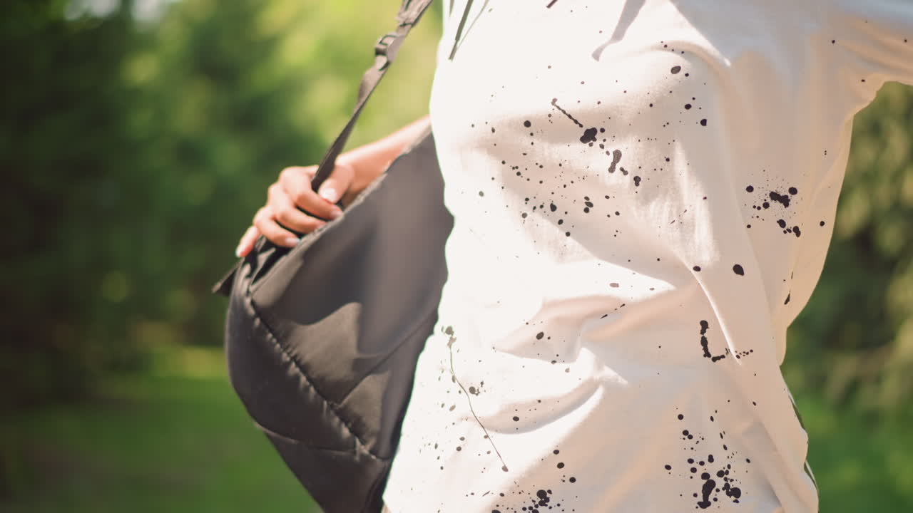 배낭을 어깨에 메고 있는 젊은 사람, 잎이 무성한 공원 산책로를 따라 걸으면서 가방 끈을 잡고 가방을 정리하는 학생의 중간 샷, 나무 사이로 따뜻한 햇빛이 쏟아지고 캐주얼한 복장을 입고 있어요.