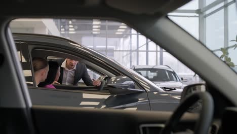 young car salesman showing to young couple new automobile at dealership salon.