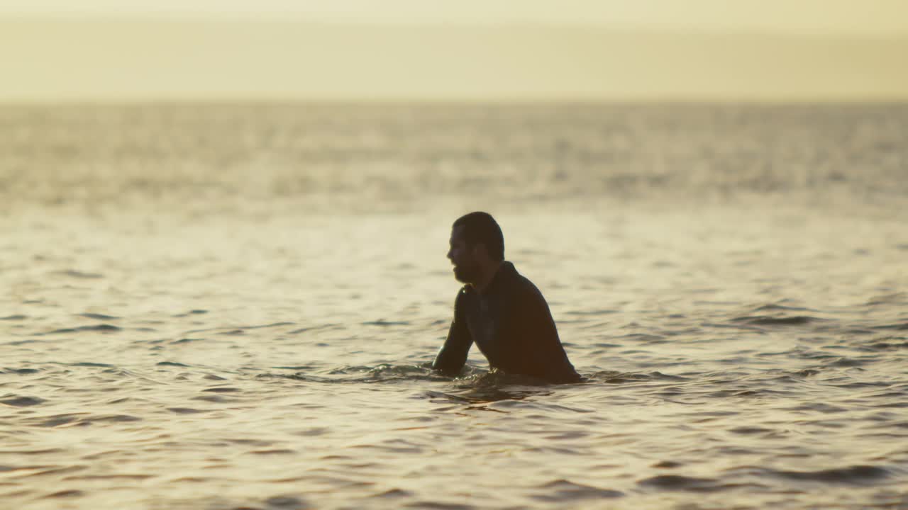 vista lateral de un surfista caucásico adulto sentado sobre una tabla de surf en el mar durante la puesta de sol 4k