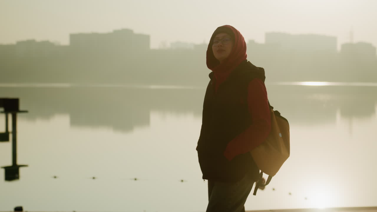 Sosok tenang berjalan-jalan di dekat danau yang tenteram saat matahari terbit. Wanita damai berjalan di tepi danau selama cahaya pagi. Siluet wanita tenang berkeliling di dekat air berkabut dengan cakrawala kota di latar belakang