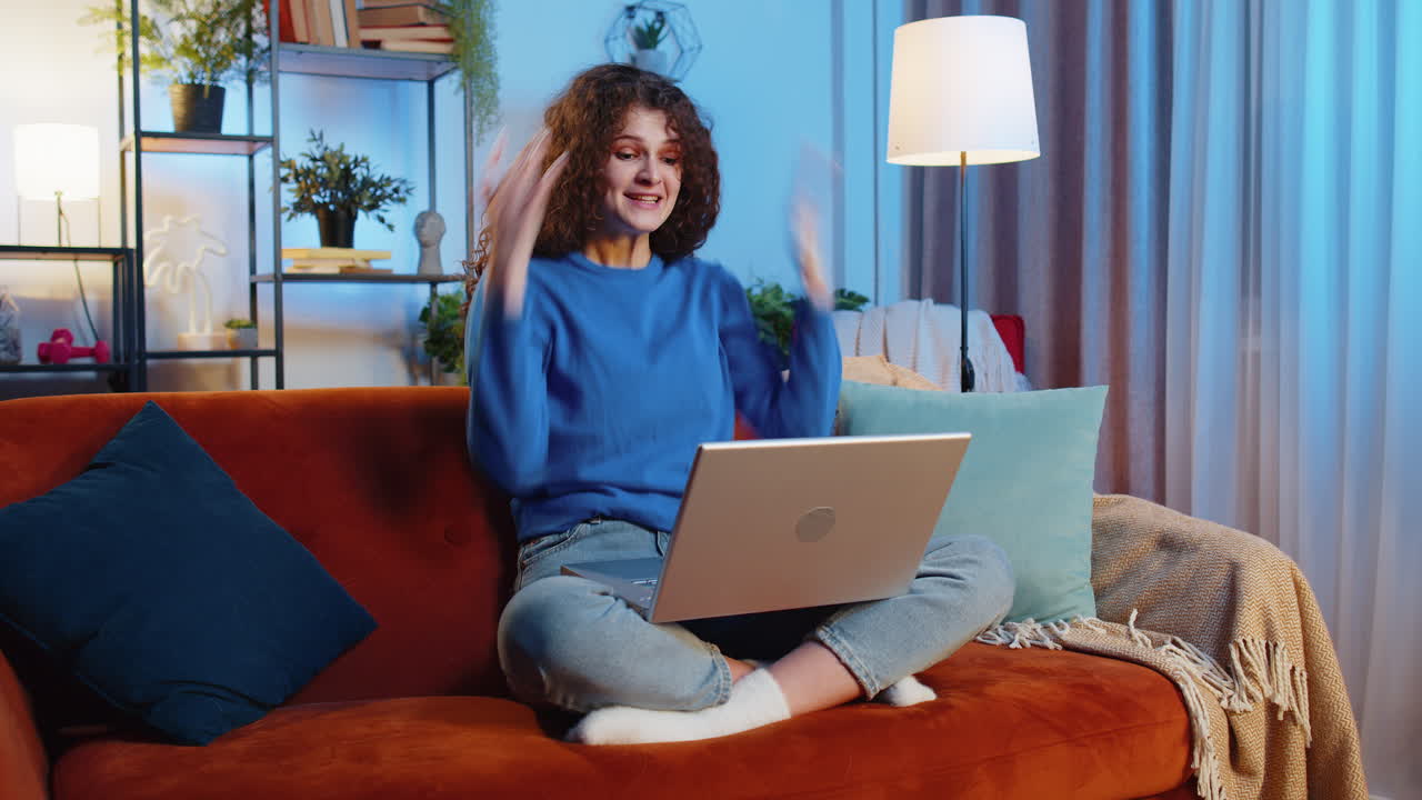 una mujer joven usando una computadora portátil y celebrando el éxito gana el baile de la victoria mientras está sentada en el sofá de casa