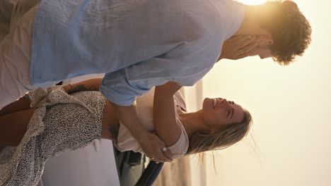 vertical video: a girl in white top holding and stroking her bearded boyfriend with curly hair on the cheek. a bearded guy in a blue shirt holds his girlfriend's hand by the shoulder in windy weather against the white convertible, a river and a yellow sky