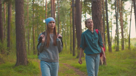 two ladies walk along forest trail surrounded by lush greenery, one playfully swings her black bag over her shoulder, while the other watches with a warm smile