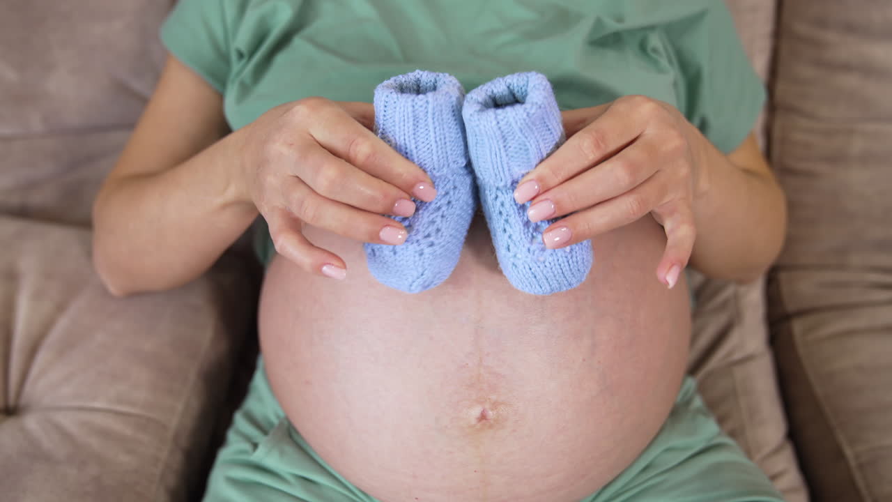 Pancione di donna incinta con scarpine da neonato. Donna incinta con accessori per bambini sul letto. Primo piano