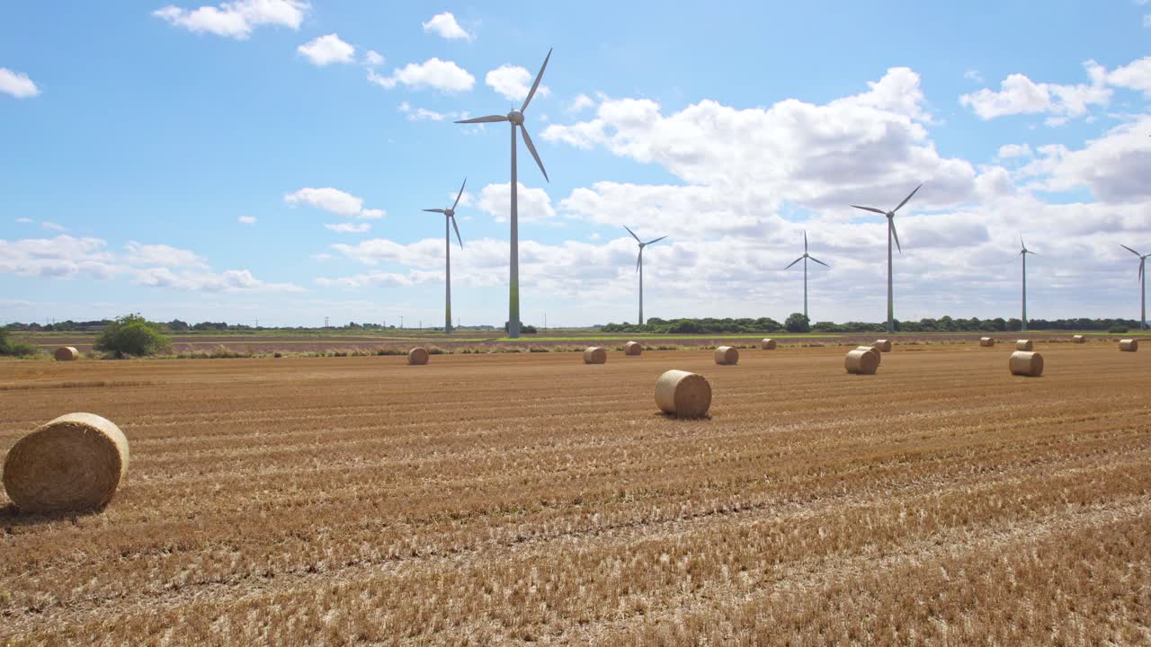 Above the Lincolnshire countryside, we see a series of wind turbines spinning in a farmer's freshly harvested field, where golden hay bales adorn the landscape