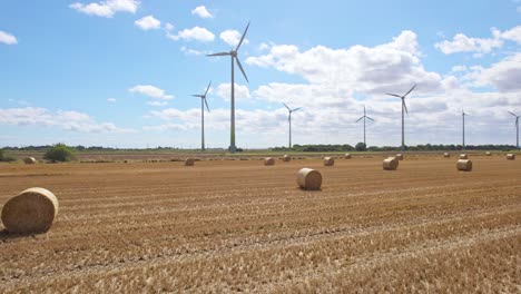 Above-the-Lincolnshire-countryside,-we-see-a-series-of-wind-turbines-spinning-in-a-farmer's-freshly-harvested-field,-where-golden-hay-bales-adorn-the-landscape