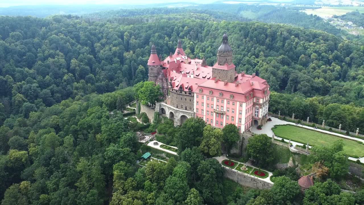 Wide aerial pullback of Ksiaz Castle in Poland, red-roofed structure among dense forest landscape