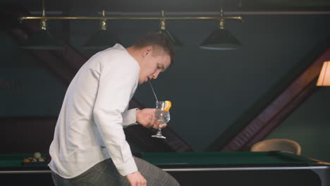 close-up of young man in white shirt and checkered trousers sitting on green pool table, holding glass of lemon drink with black straw while picking and rolling billiard ball under ambient lighting