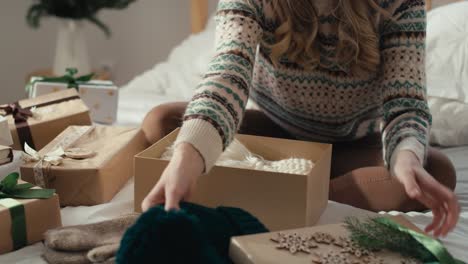 mujer caucásica sentada en la cama y empaquetando regalo de navidad en el dormitorio