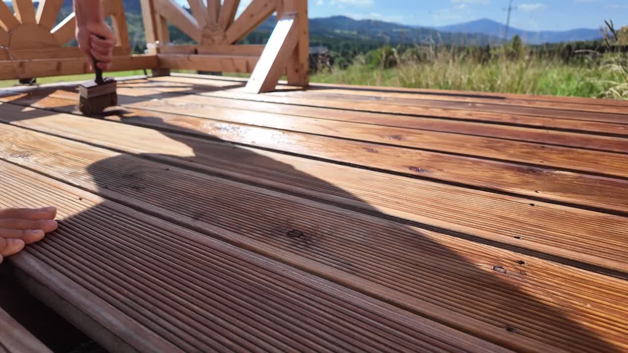 Barefooted man works to oil varnish impregnate wooden terrace boards. Decorative railing, serene sky, clouds, and mountain horizon evoke home improvement and outdoor maintenance themes - 4K