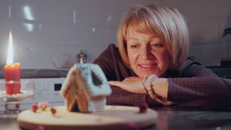 thoughtful woman rests hands on table, smiling at beautifully decorated gingerbread house, warm candlelight casts a cozy glow, creating a peaceful and nostalgic