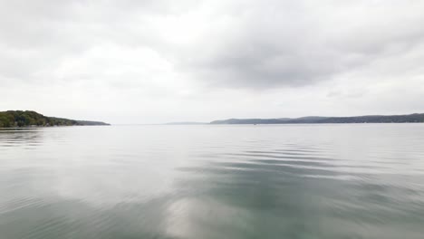 Fast-drone-shot-flying-over-Crystal-Lake-in-Beulah-Michigan-during-early-autumn-on-a-gloomy-day-with-hills-and-trees-in-the-background,-with-grey-clouds-and-a-moody-setting