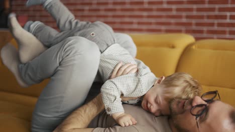 Cheerful-man-holding-jumping-toddler-boy-having-fun-together-on-sofa-in-living-room