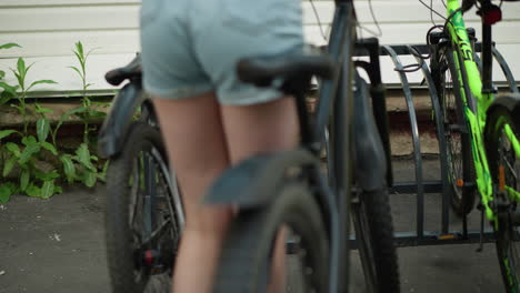 close-up rear view of young woman in jean shorts moving bicycle toward bicycle stand, positioning it among other parked bikes, background includes greenery and building