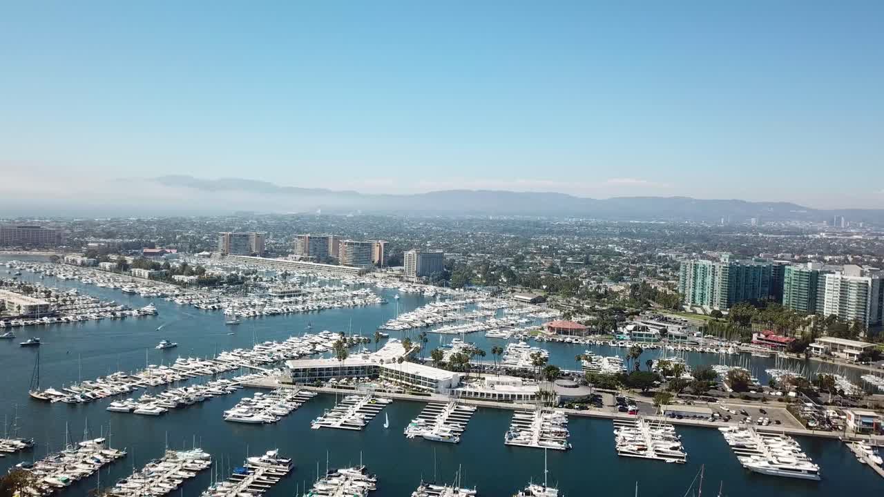 Aerial slow push-in with a pan across Marina del Rey, Los Angeles. Reveals dense marina basins filled with yachts, waterfront condos and hotels, and the Westside skyline under clear blue skies