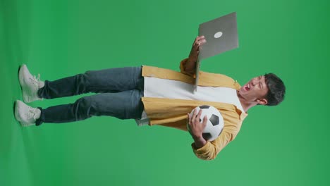 vista lateral del cuerpo completo de un niño asiático con una pelota está gritando meta alegrándose por el equipo ganador mientras anima el fútbol en una computadora portátil en el estudio de pantalla verde