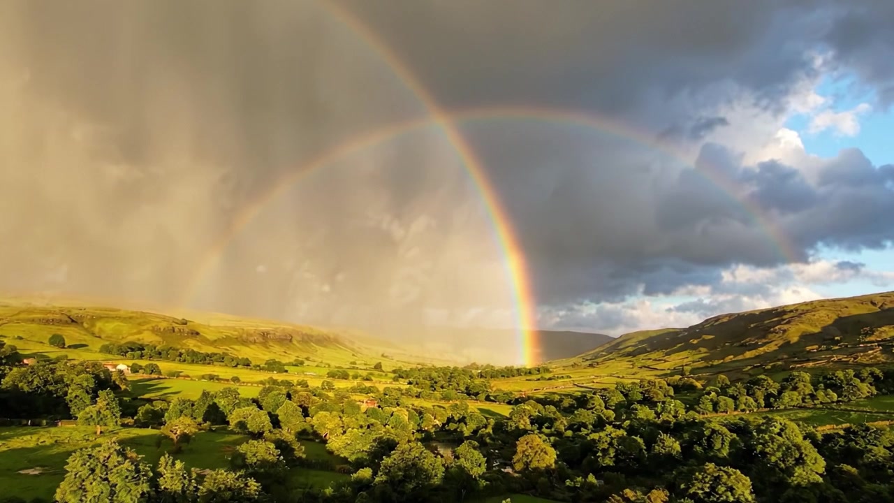 A Majestic Double Rainbow Arches Over Lush Green Hills Amidst Dramatic Skies, Showcasing Nature's Spectacular Beauty and the Calm After a Rain Shower