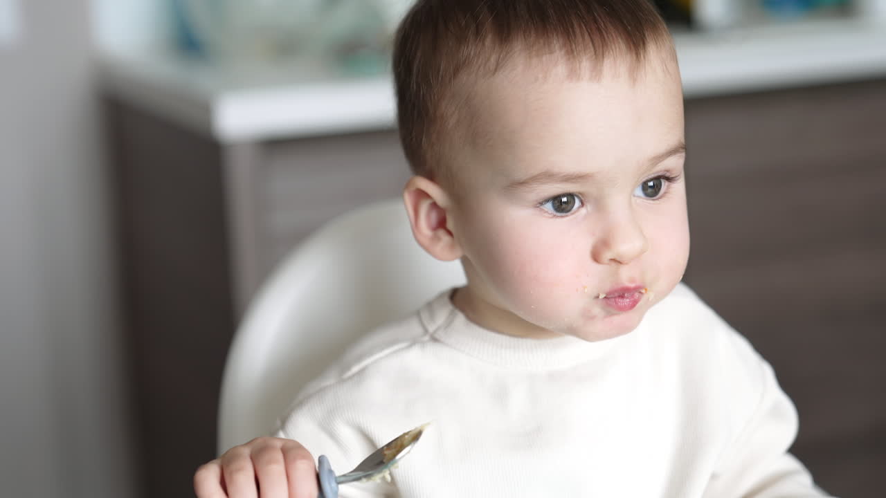 Cute Caucasian toddler slowly eating porridge from spoon. Adorable baby carefully chewing his food. Close up.