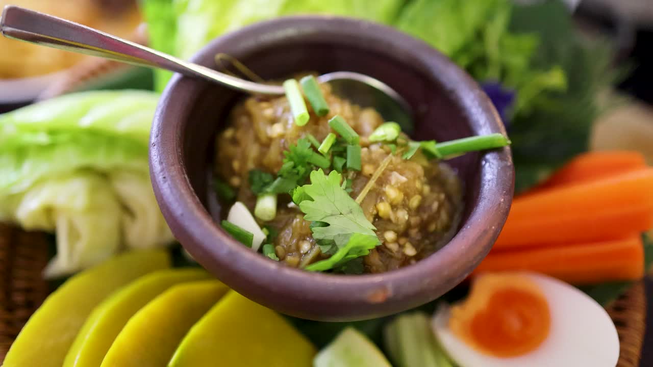 A close-up view of roasted green chili dip garnished with herbs, surrounded by assorted fresh vegetables and eggs in a traditional bamboo basket under natural lighting