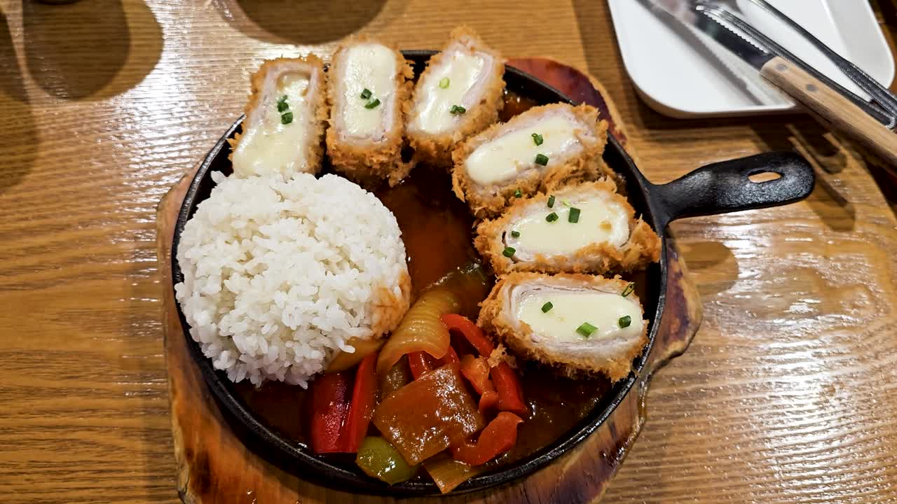 Cheesy pork cutlets arranged around steamed white rice and sautéed vegetables in savory sauce on wooden tabletop