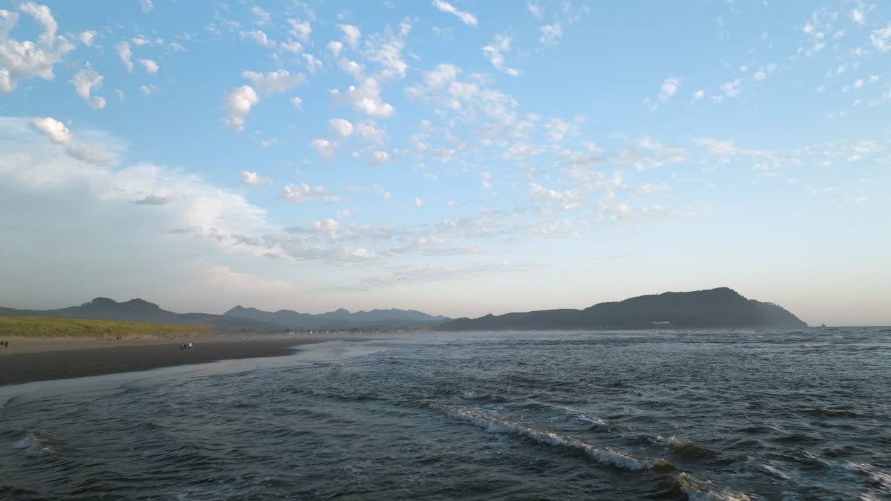 Low panoramic aerial dolly over shallow water and waves crashing on grey sand beach with mountains on horizon in distance