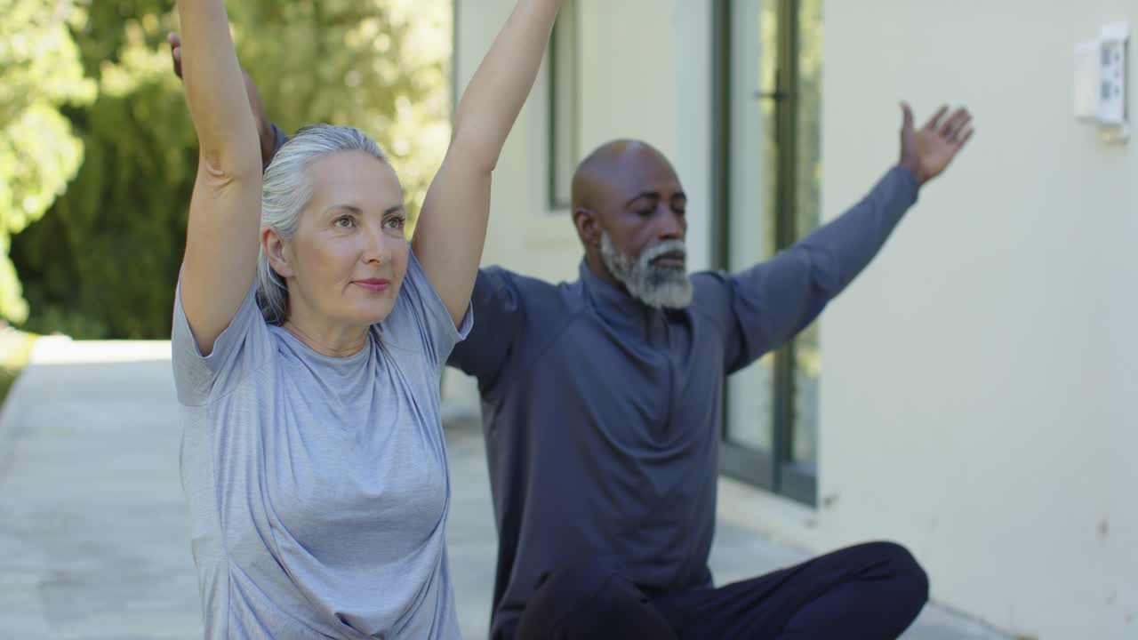 Due adulti in abbigliamento sportivo su tappetini da yoga in patio, che alzano le braccia per la calma, la donna inizia e l'uomo la imita