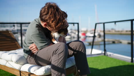 teenager boy sitting on a white deckchair is embracing his small chihuahua dog in a marina, with yachts and sailboats moored in the background
