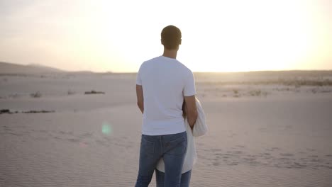 Young-couple-standing-and-hugging,-man-holding-her-woman-from-the-back-on-the-dried,-sandy-desert-watching-the-landscape-together-thoughtfully.-Overview-footage.-Lens-flares